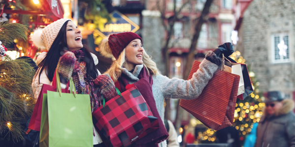 Women holiday shopping outdoors in a cold climate holding bags. Christmas tree lights shine behind them.